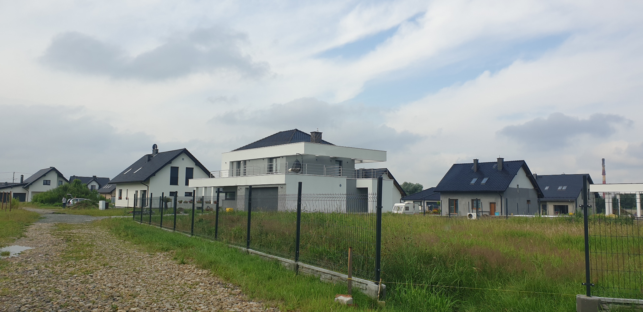 Nowoczesne domy jednorodzinne z płaskimi dachami i balkonami, widok z perspektywy drogi gruntowej, ogrodzenie panelowe na pierwszym planie.