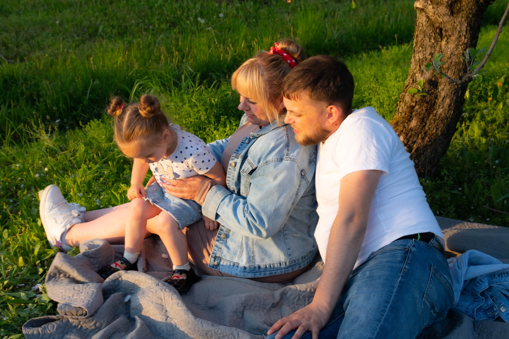 Rodzina siedząca na kocu w trawie pod drzewem w ciepłym świetle zachodzącego słońca. Córka siedzi na kolanach mamy, ubrana w dżinsową spódniczkę i bluzkę w kropki.