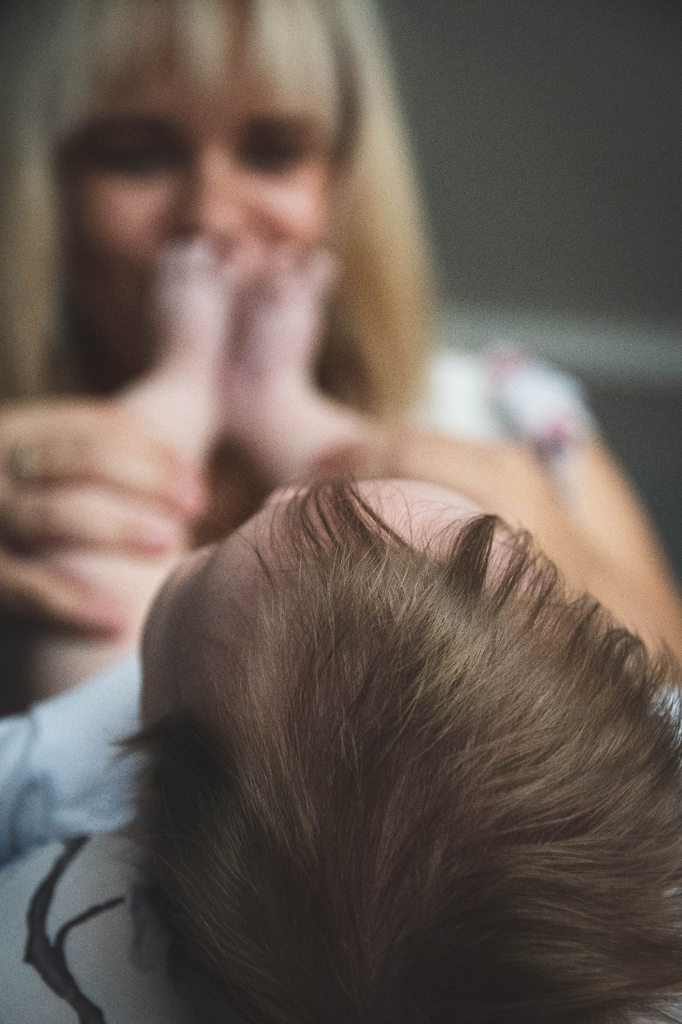 Głowa niemowlęcia z widocznymi włosami, w tle rozmyta twarz kobiety trzymającej stopy dziecka.
