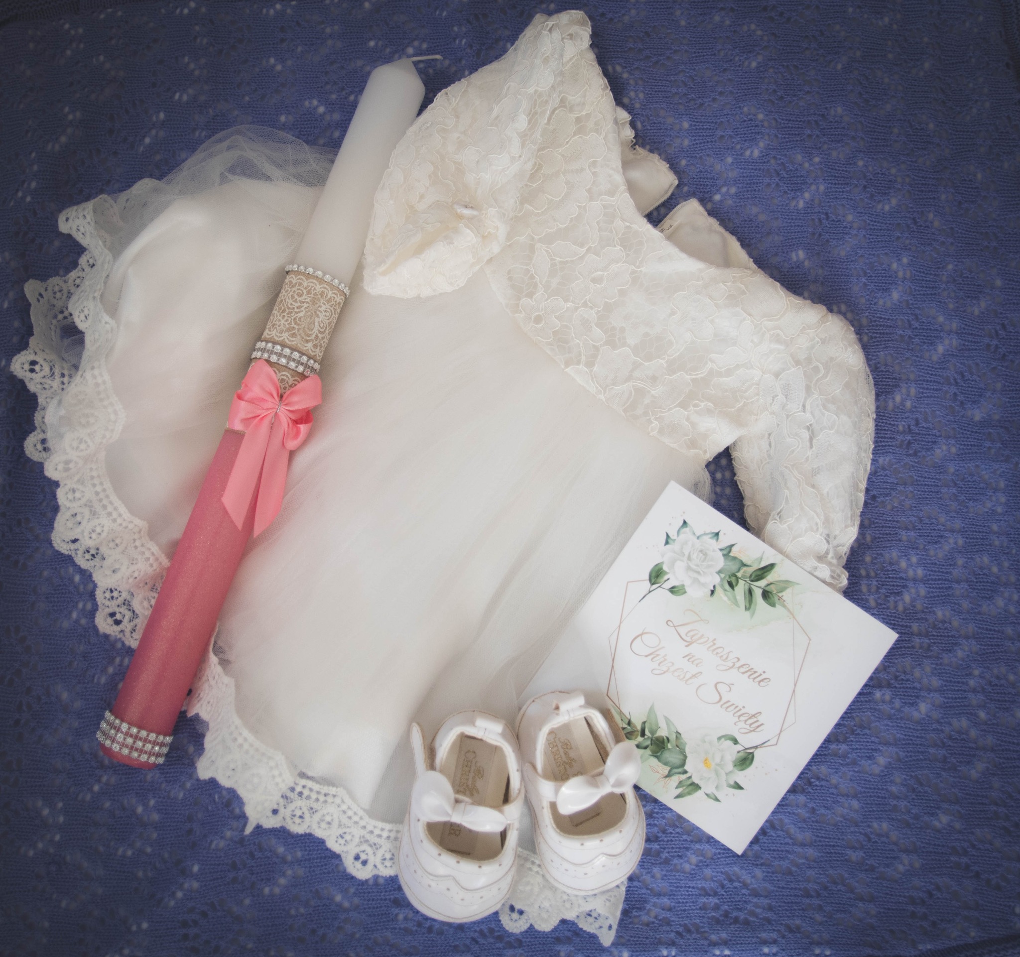 Komplet ubranek do chrztu: biała sukienka z koronkowymi rękawami, świeca z różową wstążką, zaproszenie na chrzest i białe buciki na tle niebieskiego koca.