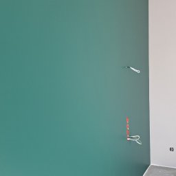 Tar-Bud - Świeżo pomalowana ściana na zielono w narożniku pomieszczenia, widoczne wystające kable elektryczne przed montażem osprzętu, surowa podłoga w trakcie prac wykończeniowych.
