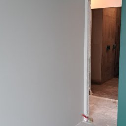 Tar-Bud - Świeżo pomalowane na szaro ściany i podłoga w trakcie prac wykończeniowych, widoczna szpachelka i pędzel przy listwie przypodłogowej, w tle surowa łazienka z widocznymi przyłączami.