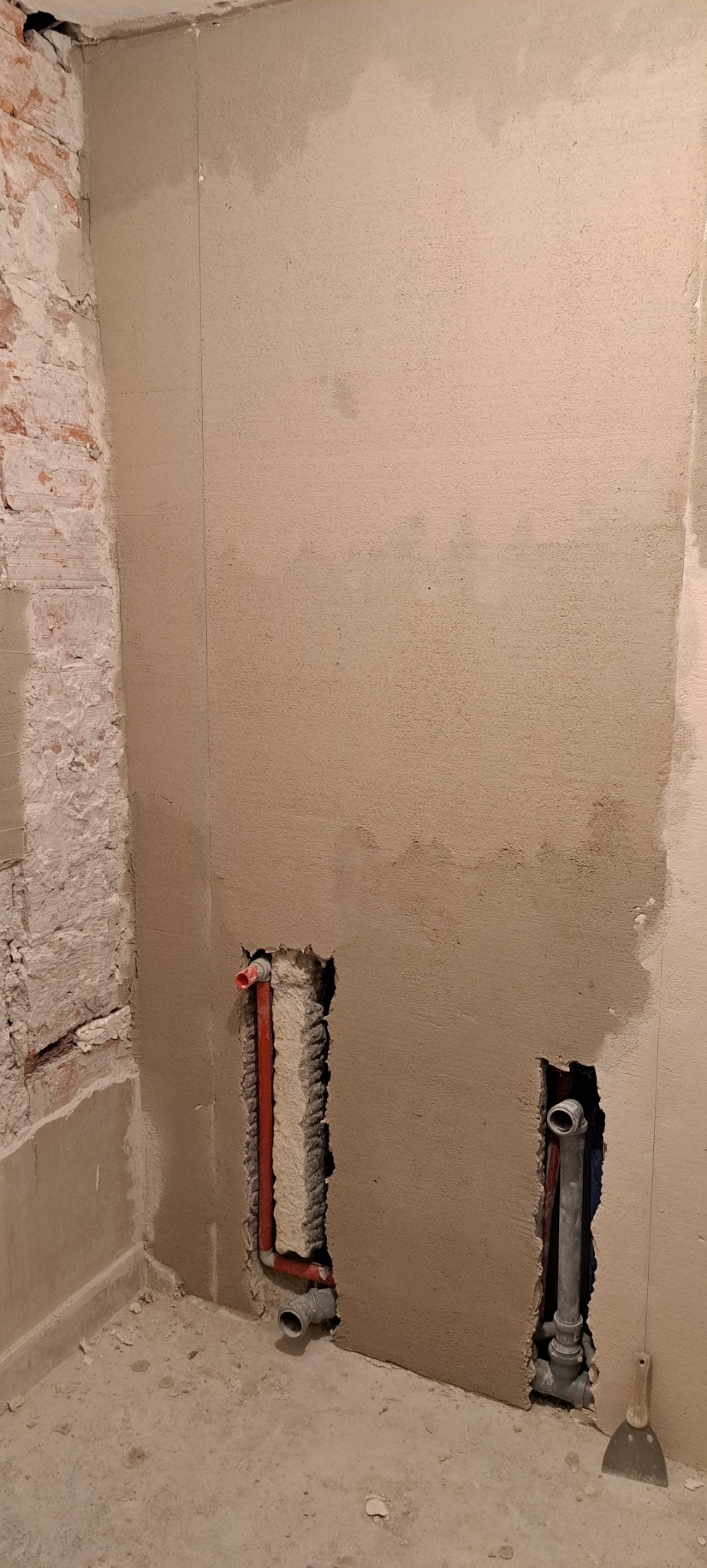 Ściana w trakcie tynkowania z widocznymi instalacjami hydraulicznymi i elektrycznymi w przygotowanych otworach, fragment odsłoniętej cegły, narzędzie szpachelka przy ścianie.