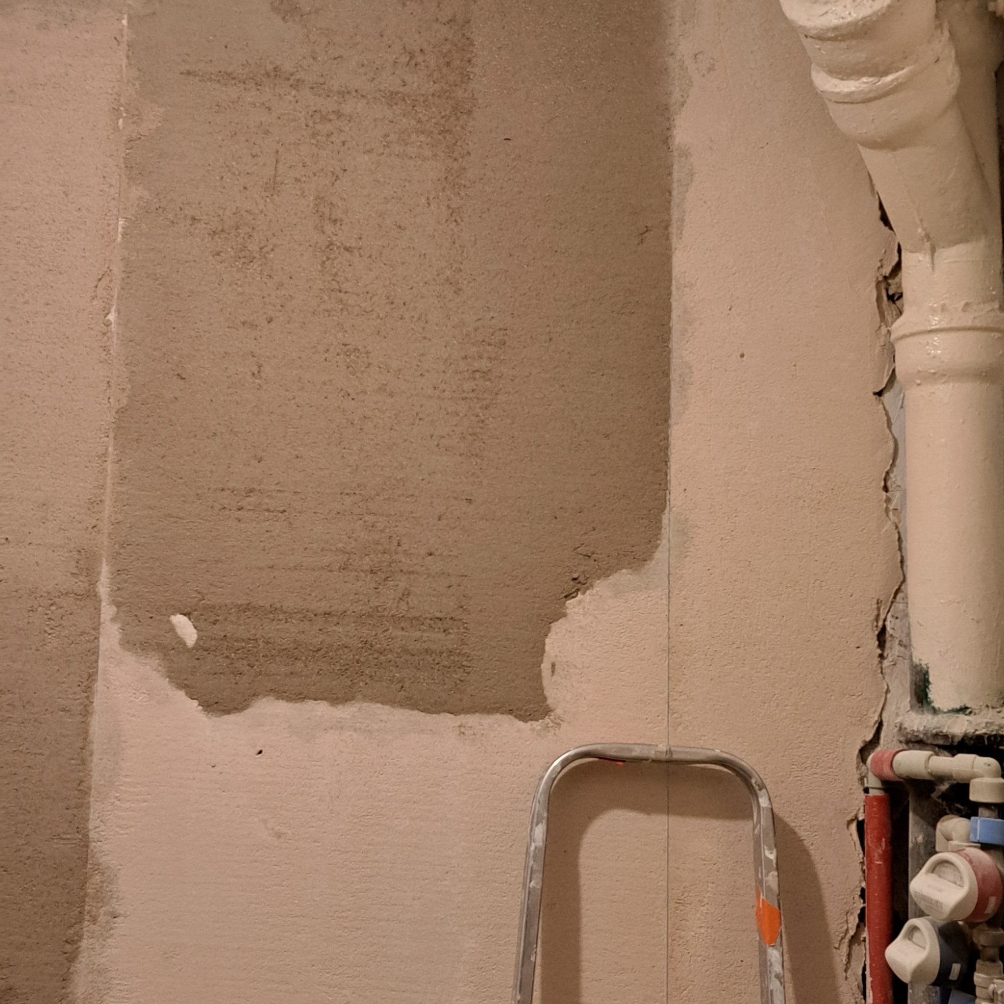 Fragment ściany w trakcie szpachlowania, widoczne rury instalacyjne i fragment drabiny malarskiej.