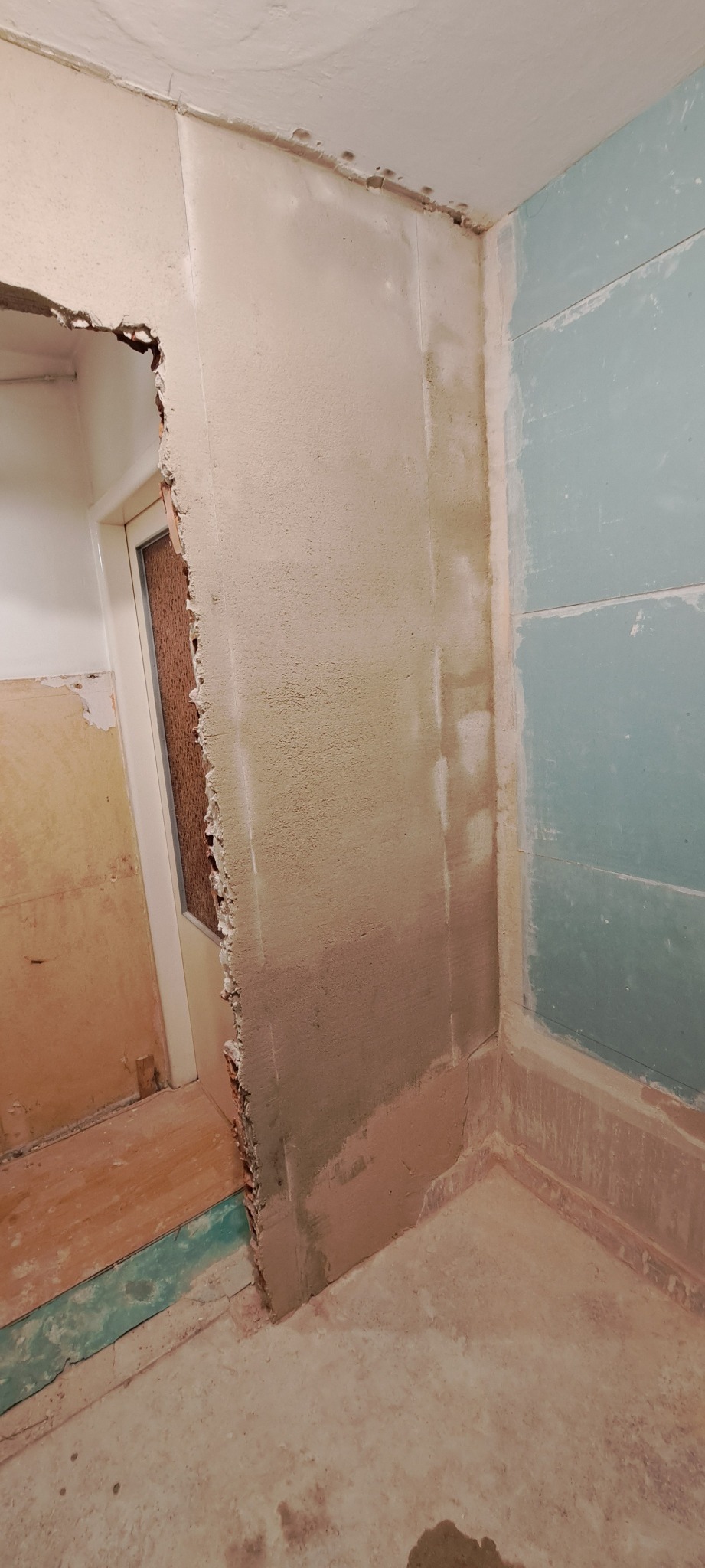 Fragment łazienki w trakcie prac przygotowawczych: usunięte płytki, częściowo otynkowane ściany, widoczne nierówności i ślady po kleju, surowy stan przed dalszym wykończeniem.