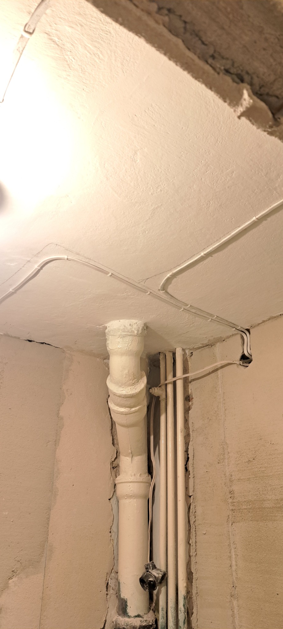 Rury kanalizacyjne i instalacja elektryczna poprowadzone po ścianach i suficie w trakcie remontu łazienki. Widoczne ślady po starych powłokach malarskich i uszkodzenia tynku.