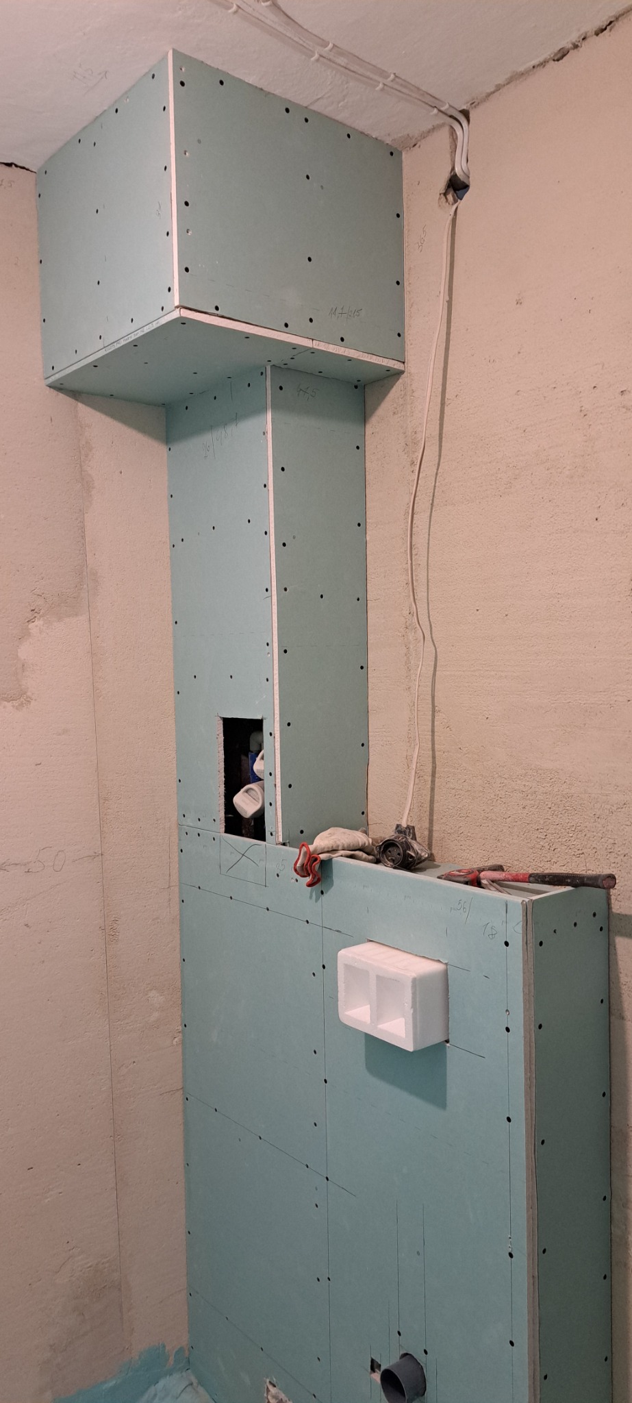 Kąt w łazience w trakcie zabudowy stelaża WC płytami gipsowo-kartonowymi; widoczne narzędzia, instalacja elektryczna i fragment ściany z surowym tynkiem.