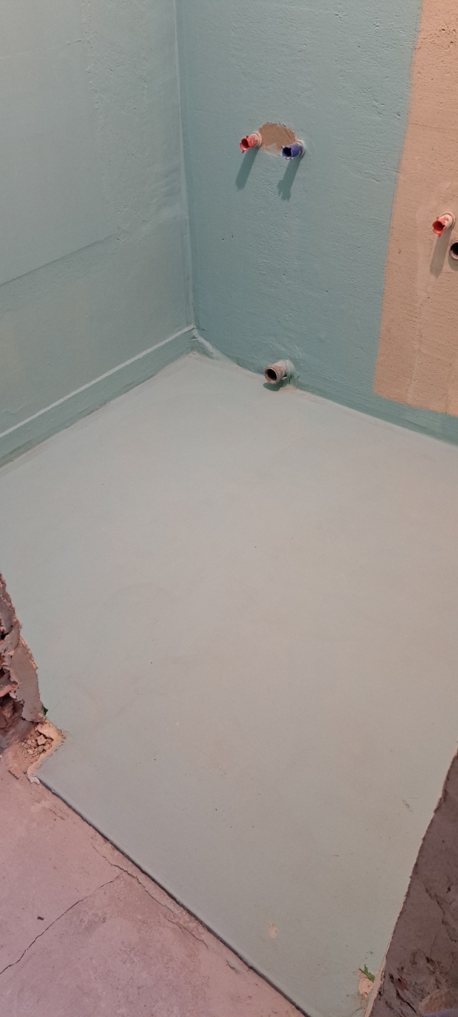 Przygotowanie podłoża w łazience: hydroizolacja w kolorze jasnej zieleni na ścianach i podłodze, widoczne wystające rury instalacji wodnej, surowy stan wykończenia przy podłodze.