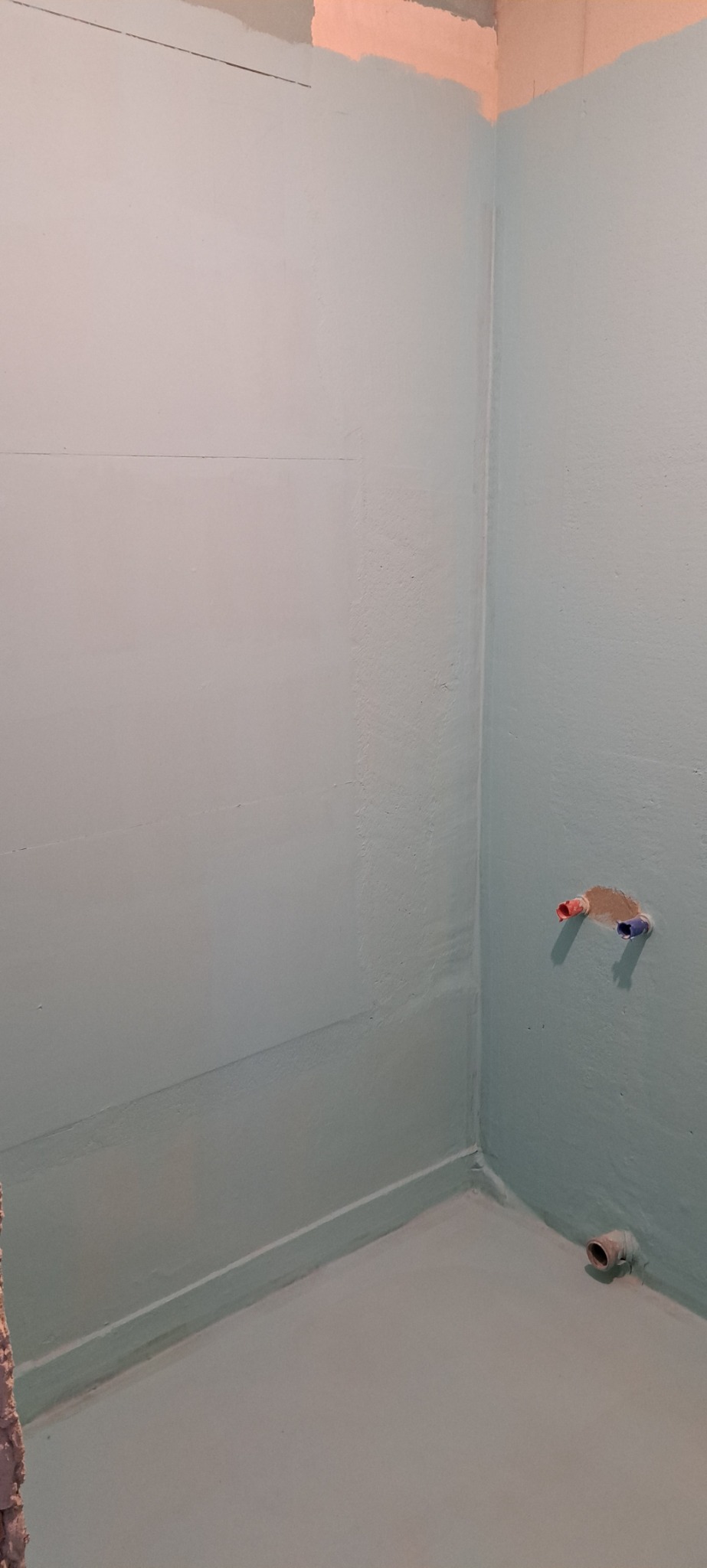 Świeżo pomalowane wnętrze łazienki na jednolity, jasny, pastelowy odcień niebieskiego, widoczne wystające rury instalacji wodnej i kanalizacyjnej, przygotowane do dalszych prac wykończeniowych.