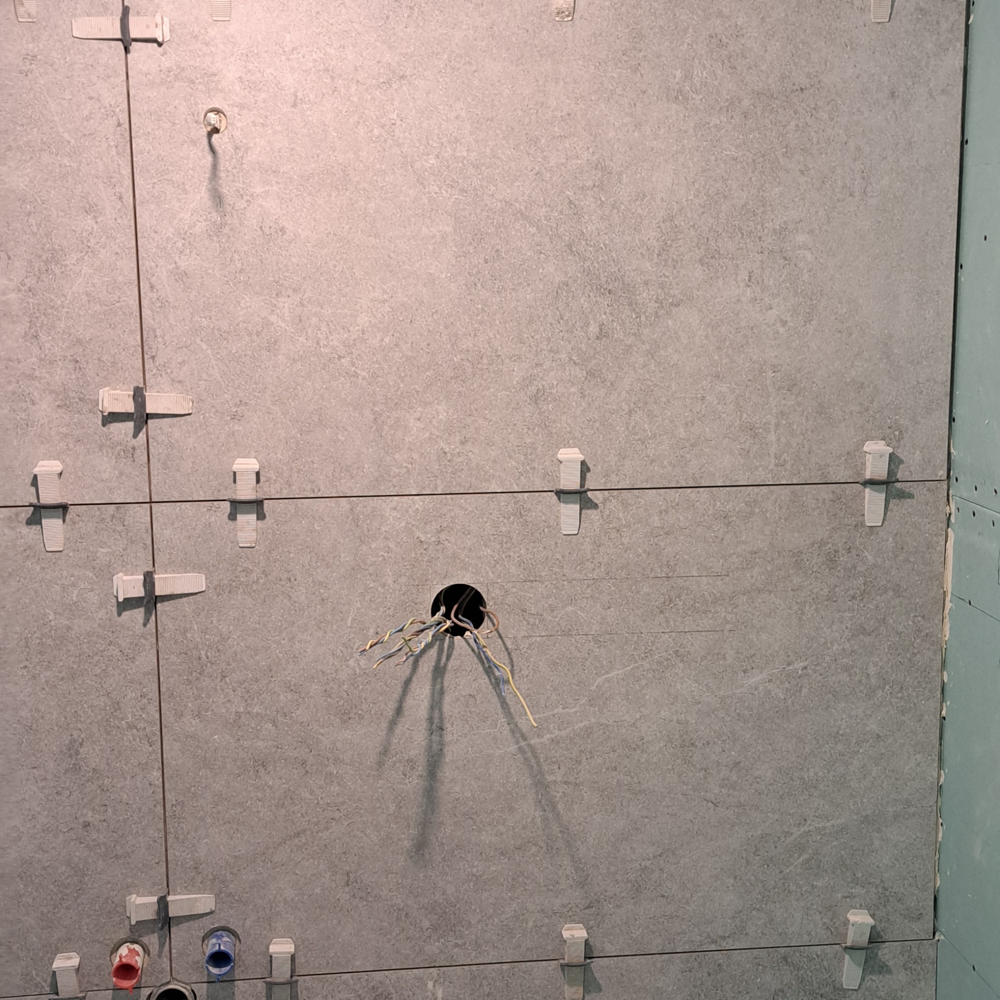 Szare płytki ścienne w trakcie montażu, z widocznymi krzyżykami dystansowymi, otworem na instalację elektryczną z wystającymi przewodami i podłączeniami hydraulicznymi w dolnej części ściany.