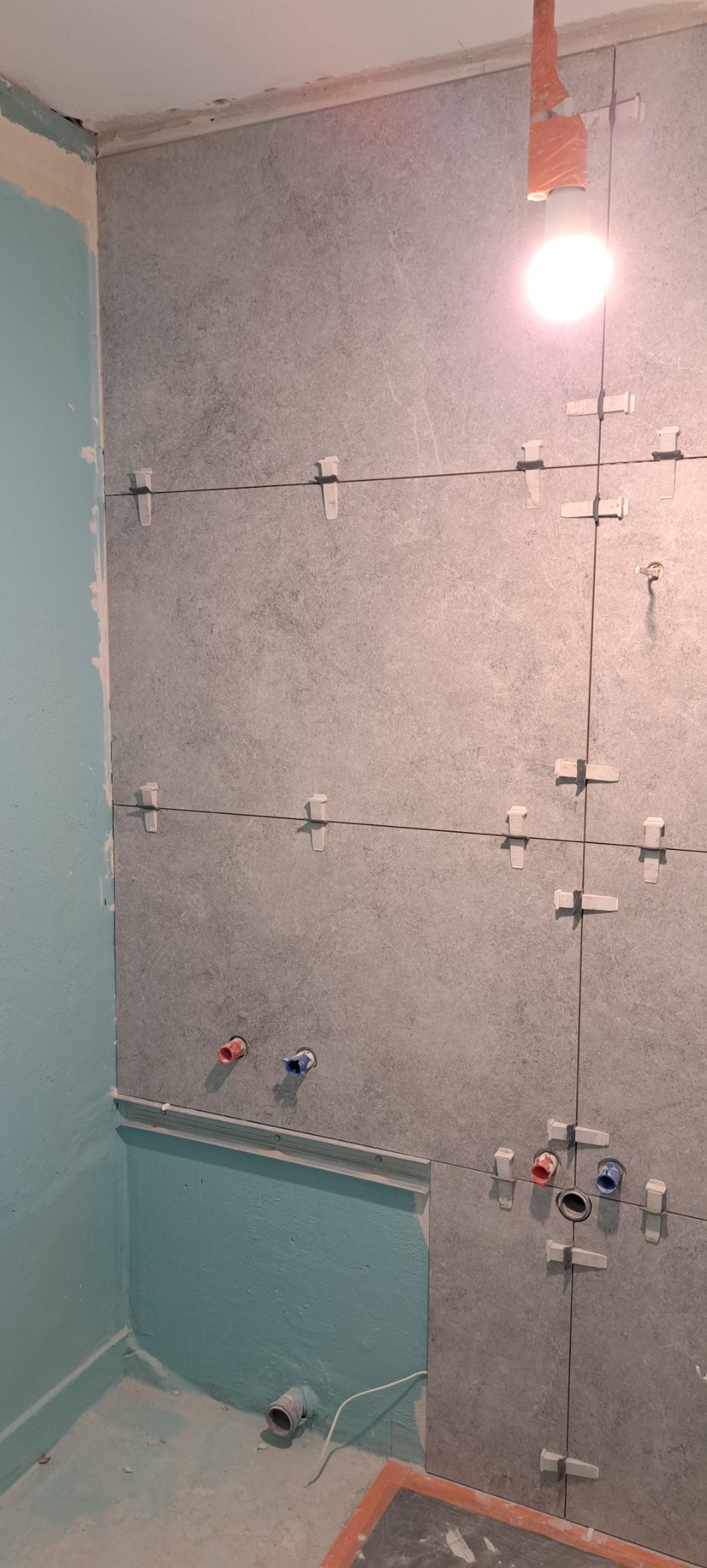 Ściana łazienki w trakcie układania szarych płytek z użyciem krzyżyków dystansowych, widoczne rury doprowadzające wodę i fragment podłogi z odpływem