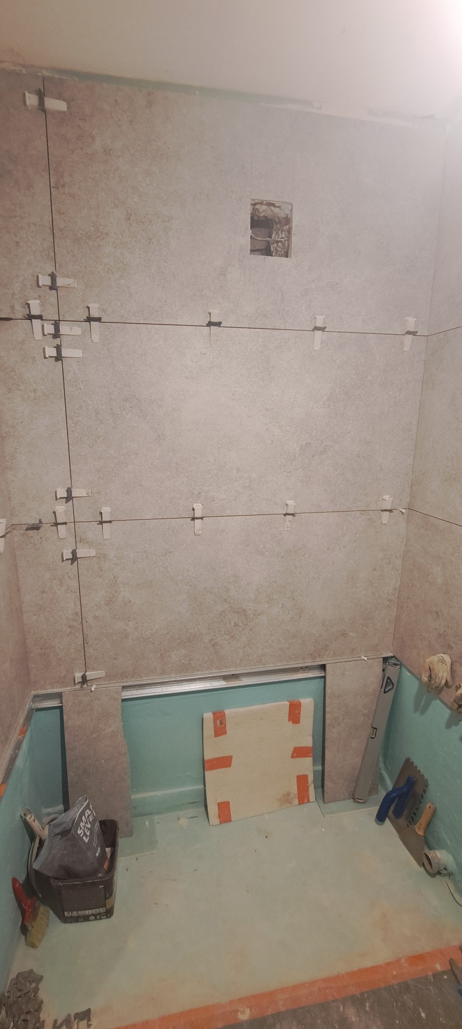 Proces układania płytek w łazience, widoczne płytki na ścianach z krzyżykami dystansowymi, poziomica, wiadro z zaprawą, oraz przygotowane podłoże pod płytki.