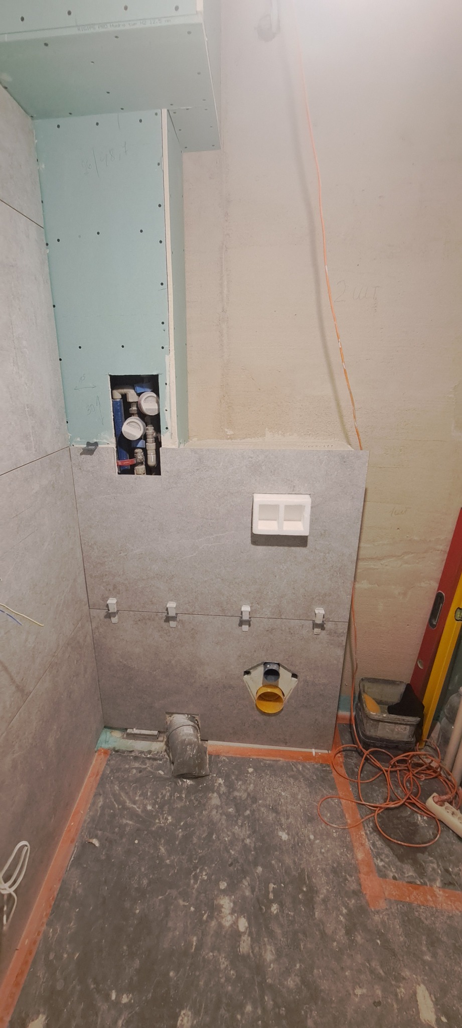 Łazienka w trakcie remontu: szare płytki częściowo położone, widoczne elementy instalacji wodnej i kanalizacyjnej, płyta gipsowo-kartonowa z wyciętym otworem na rury, poziomica i narzędzia w rogu.