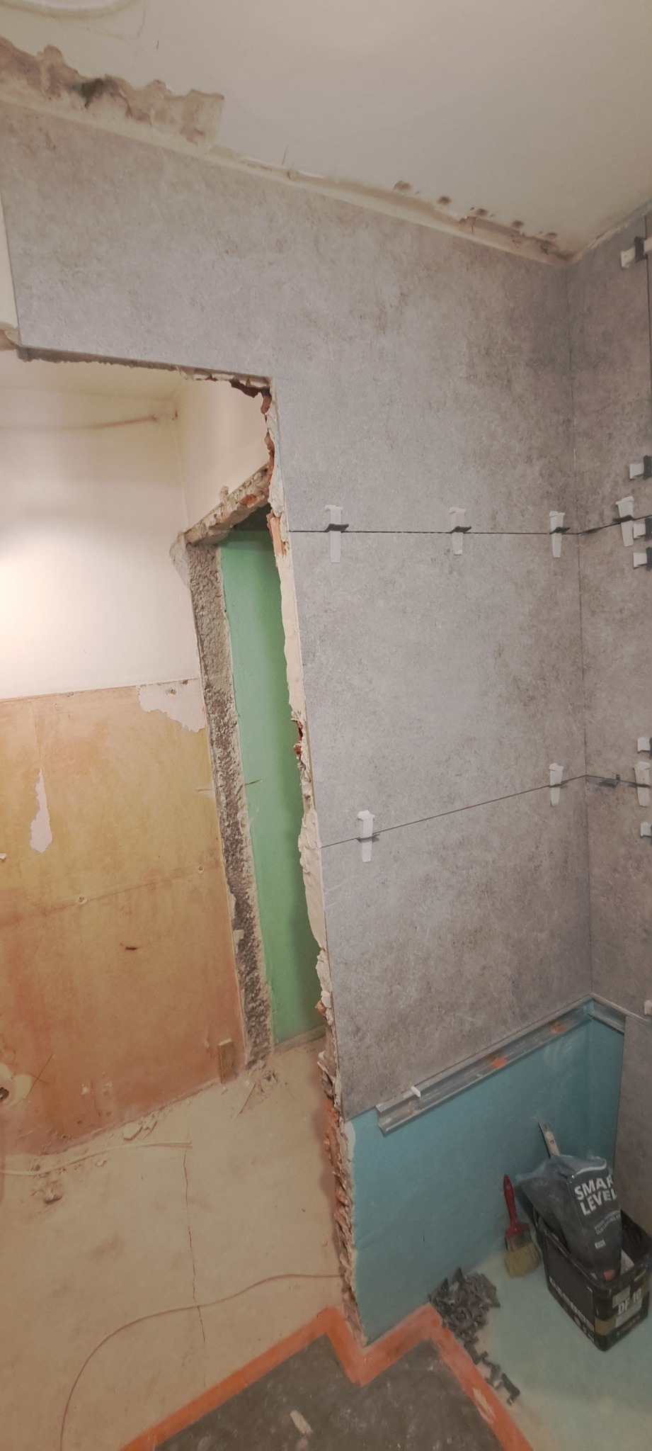 Fragment ściany łazienki w trakcie układania szarych płytek, widoczne krzyżyki dystansowe, listwa pod wanną i narzędzia w pojemniku.