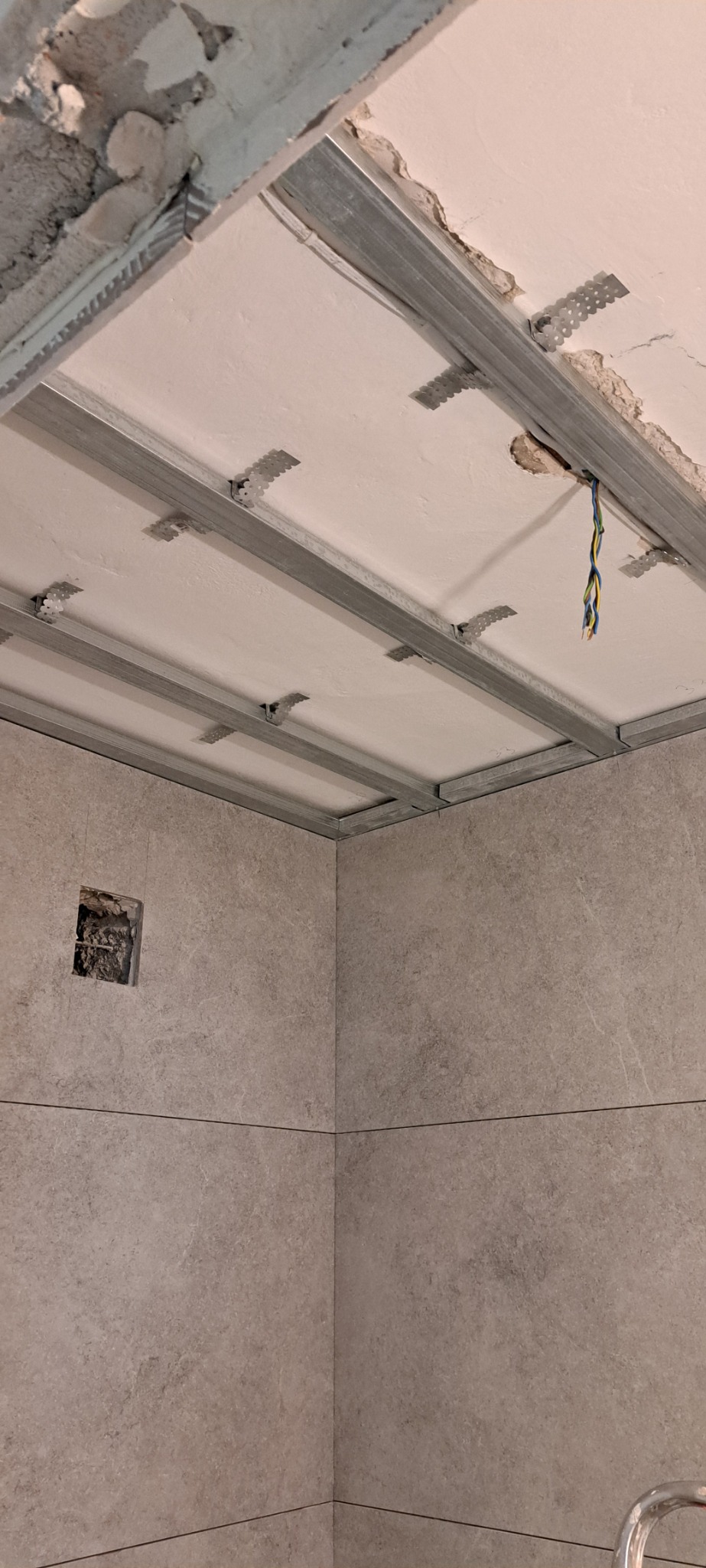 Montaż sufitu podwieszanego z metalowej konstrukcji w łazience z szarymi płytkami, widoczne wystające kable i otwór w ścianie.