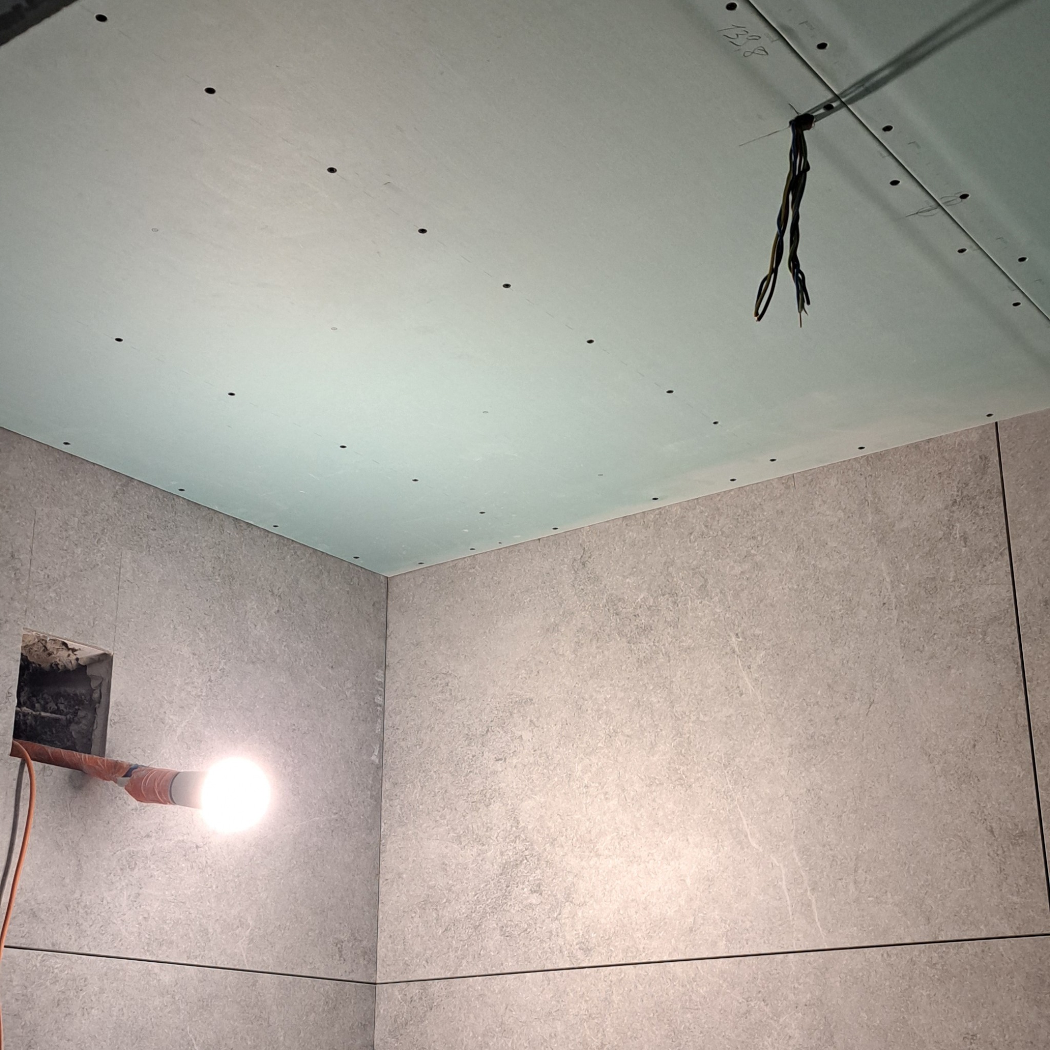 Wnętrze łazienki w trakcie wykańczania: szare płytki imitujące kamień na ścianach, sufit podwieszany z widocznymi otworami na oświetlenie, wystające przewody elektryczne i tymczasowe oświetlenie...