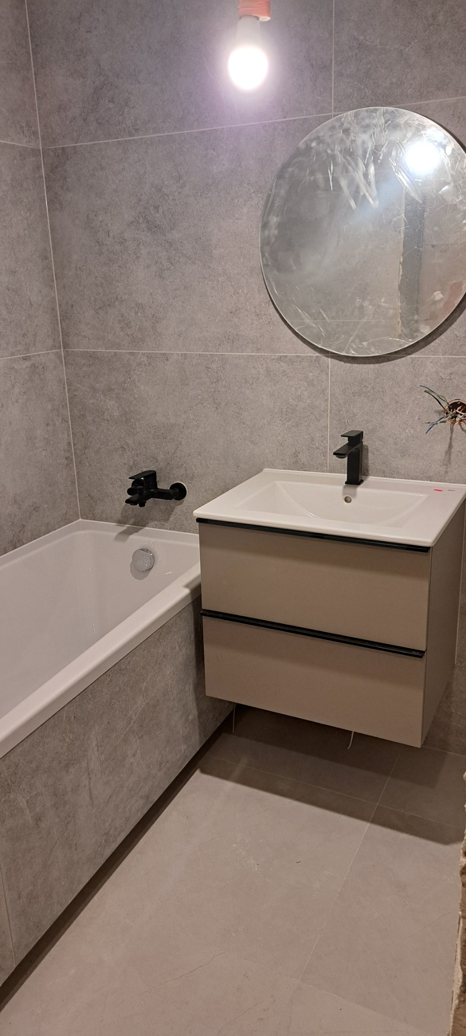 Wykończona łazienka z szarymi płytkami, białą wanną obłożoną szarymi płytkami, beżową szafką pod umywalkę z czarną armaturą i okrągłym lustrem, widoczne wystające kable elektryczne.