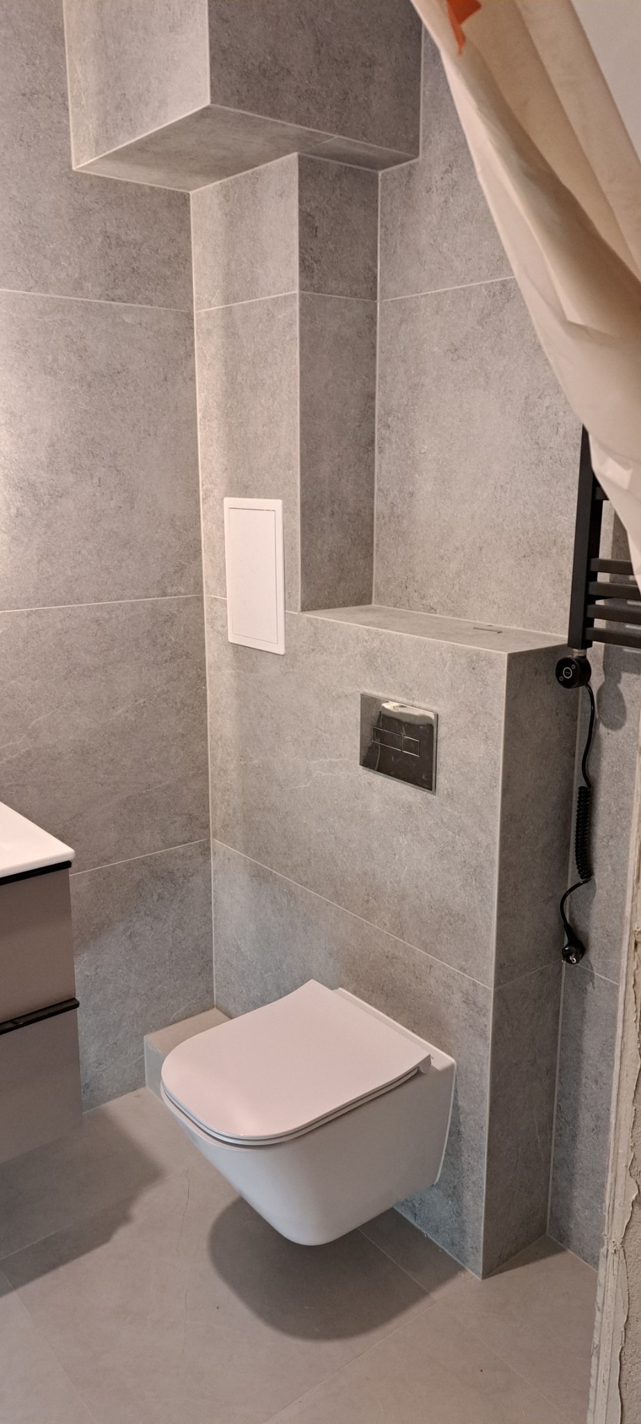 Nowoczesna łazienka z wiszącą toaletą, szare płytki imitujące kamień, chromowany przycisk spłukiwania, ukryty rewizyjny właz i grzejnik drabinkowy z kablem spiralnym.