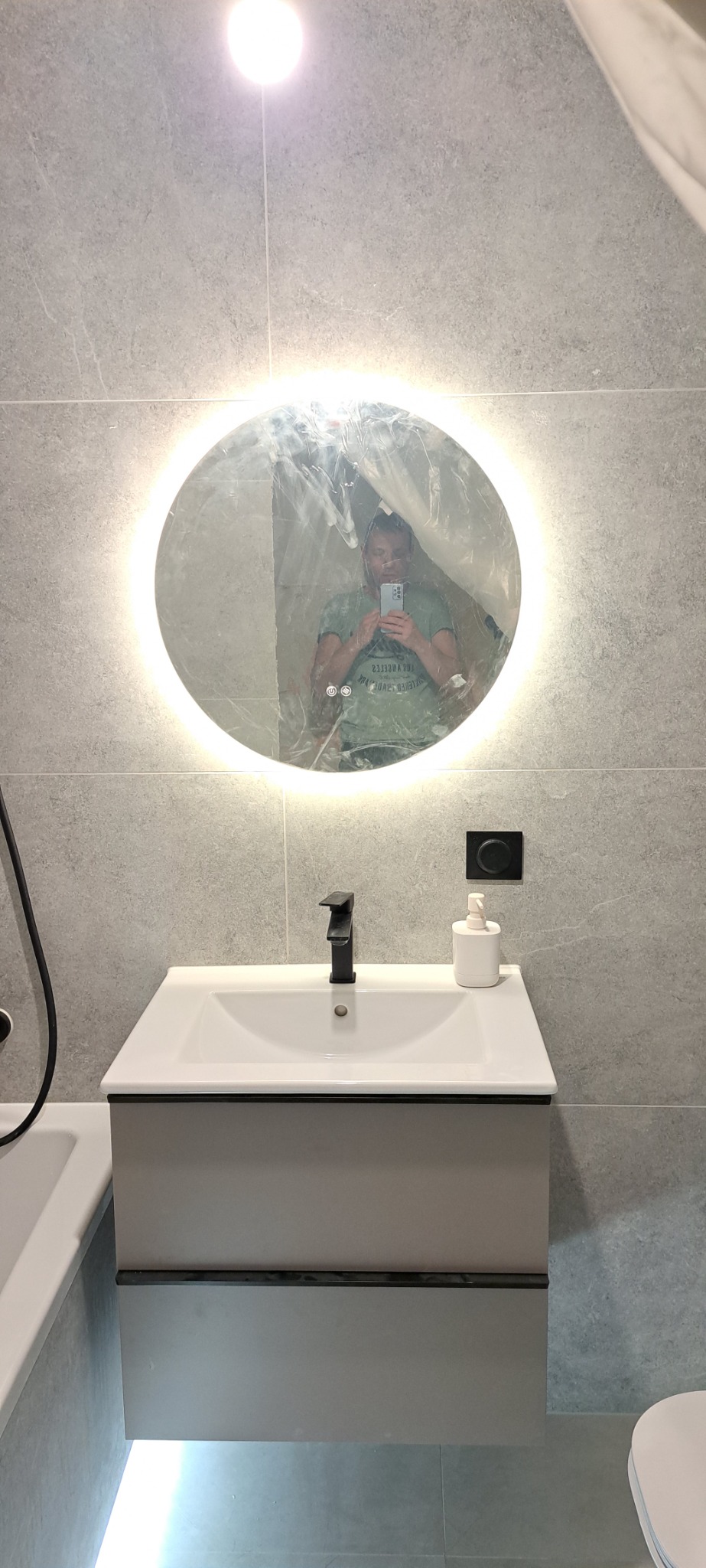 Nowoczesna łazienka w szarych tonacjach z okrągłym lustrem LED i białą umywalką z czarną armaturą, uchwycona w trakcie remontu z widoczną folią ochronną na lustrze i odbiciem osoby robiącej zdjęcie.