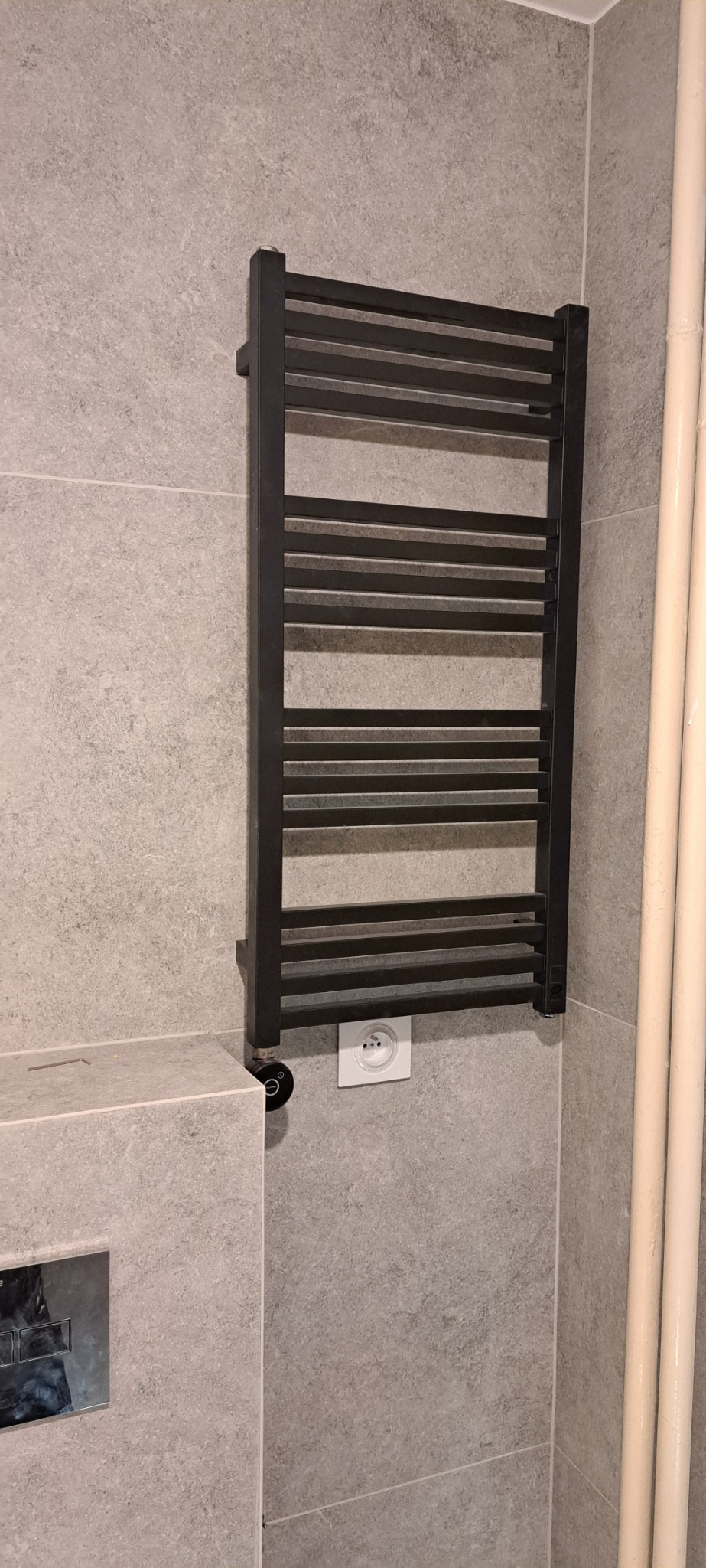Czarny, drabinkowy grzejnik łazienkowy zamontowany na szarej, kamiennej ścianie, widoczny fragment instalacji wodnej i gniazdko elektryczne.