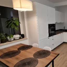 Jasna kuchnia z białymi szafkami i czarnymi akcentami, widok na stół z dekoracyjnymi podkładkami i rośliny w oknie.