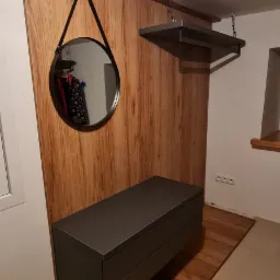 Nowoczesna zabudowa przedpokoju z panelem drewnopodobnym, szafką z szufladami w kolorze grafitowym, okrągłym lustrem na pasku i półką pod sufitem