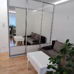 Nowoczesna szafa wnękowa z przesuwnymi lustrzanymi drzwiami w jasnym pokoju, odbijająca sofę i stolik kawowy, z rośliną doniczkową w rogu.