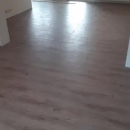 Ułożone panele podłogowe w jasnym odcieniu drewna w pustym pokoju z białymi ścianami i oknem balkonowym, widoczne elementy instalacji grzewczej i torba na parapecie.