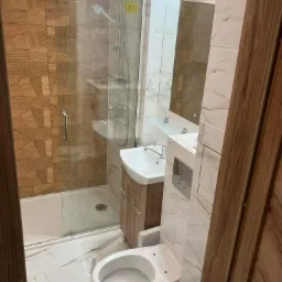 Nowoczesna łazienka z kabiną prysznicową wyłożoną płytkami imitującymi drewno, białą umywalką z szafką w odcieniach drewna oraz toaletą. Na podłodze i ścianach białe płytki z marmurkowym wzorem.