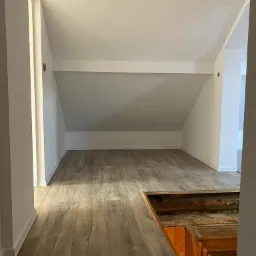 Poddasze z wykończoną podłogą z paneli imitujących drewno, białe ściany i sufit, widoczne schody w trakcie prac wykończeniowych.