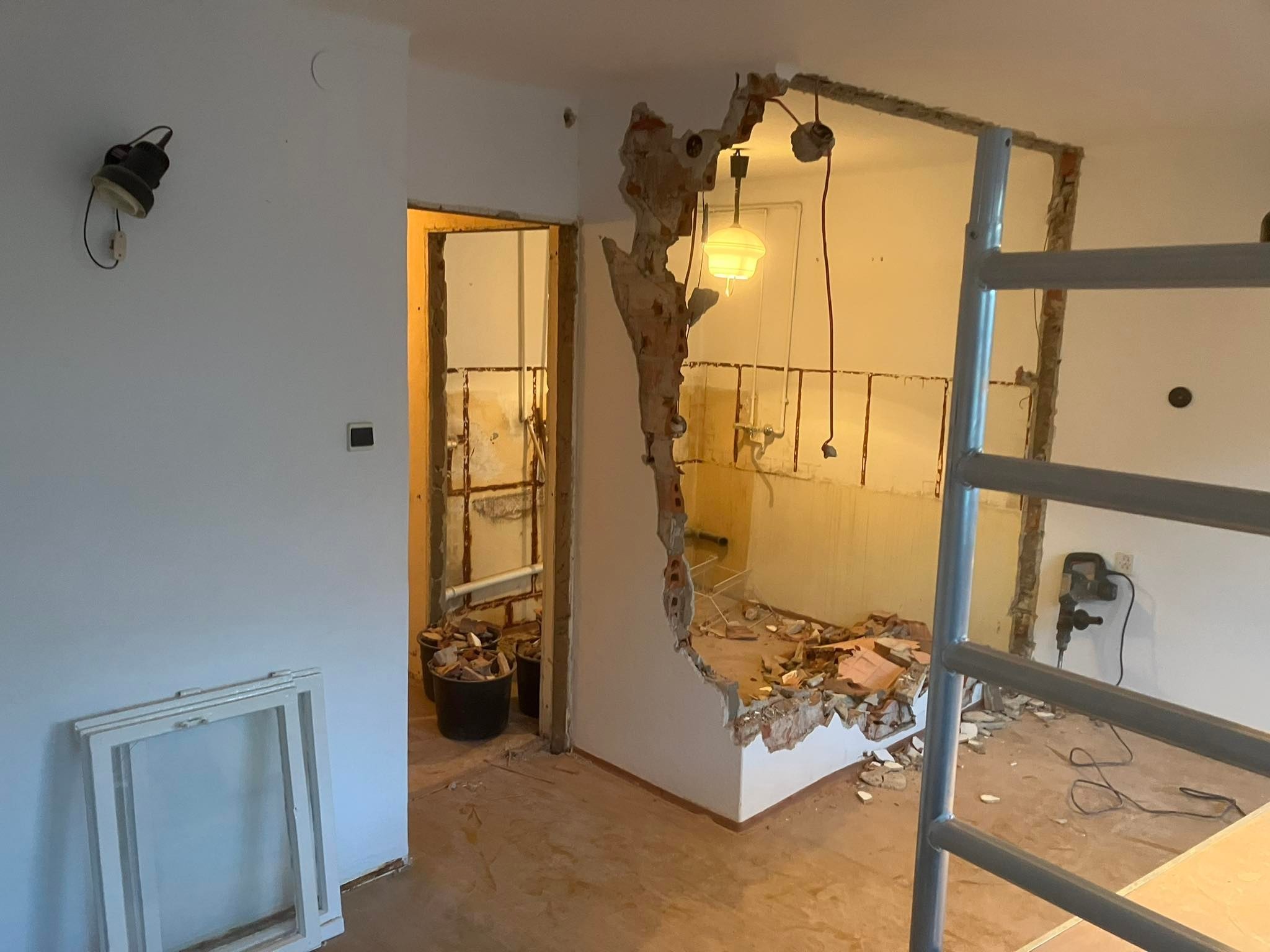 Demontaż ściany działowej w trakcie prac remontowych, widoczne fragmenty instalacji, gruz w wiadrach, stojące okno i drabina, wisząca lampa