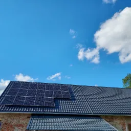Panele słoneczne na ciemnym dachu krytym dachówką, z widoczną ceglaną ścianą budynku i błękitnym niebem z chmurami w tle.