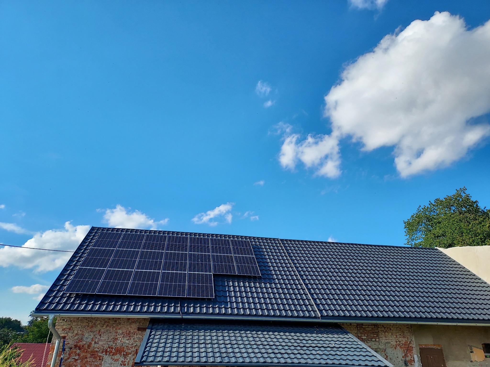 Panele słoneczne na ciemnym dachu krytym dachówką, z widoczną ceglaną ścianą budynku i błękitnym niebem z chmurami w tle.
