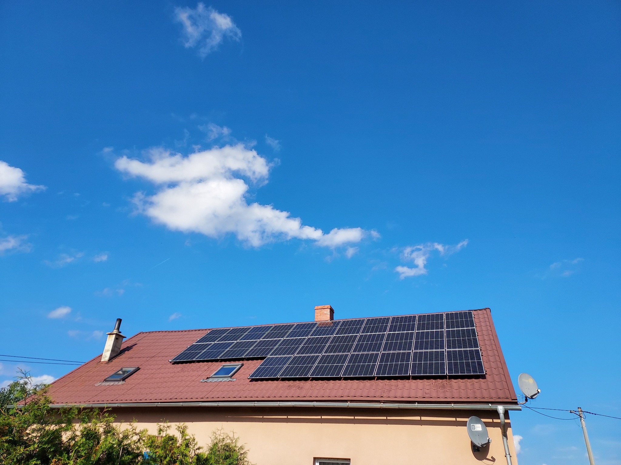 Dom jednorodzinny z brązowym dachem pokrytym panelami słonecznymi, widoczne dwa okna dachowe, komin i anteny satelitarne, na tle błękitnego nieba z białymi chmurami.