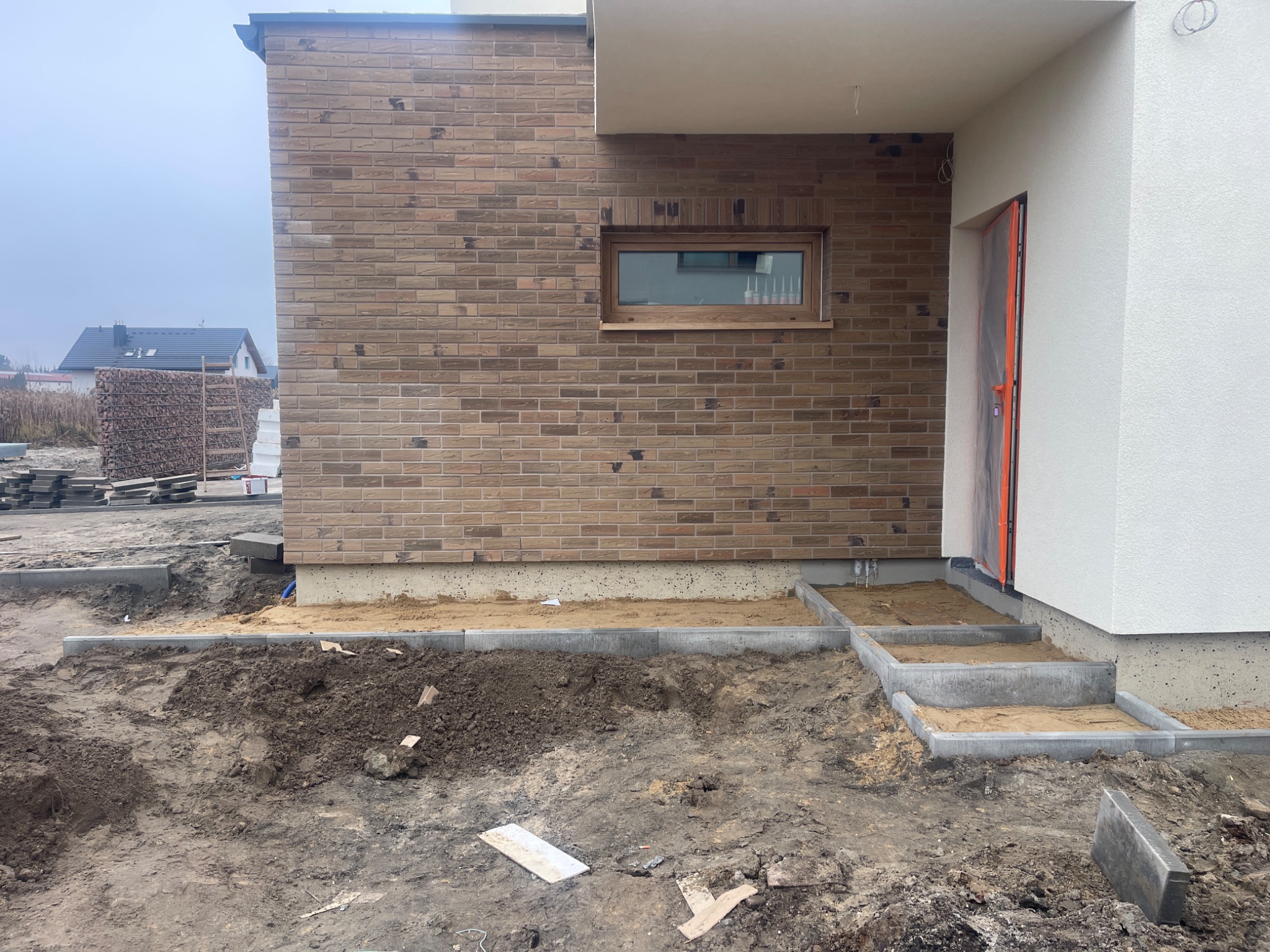 Nowoczesny dom w trakcie wykończenia: ceglana elewacja z oknem, biały fragment ściany z drzwiami zabezpieczonymi folią, ułożone schody z kostki brukowej, teren przygotowany pod kostkę.