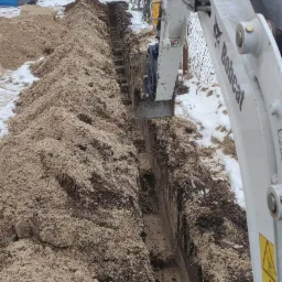 Wąski wykop w piaszczystej glebie z widoczną łyżką koparki Bobcat, częściowo pokryty śniegiem, na tle zabudowań mieszkalnych i ogrodzenia.