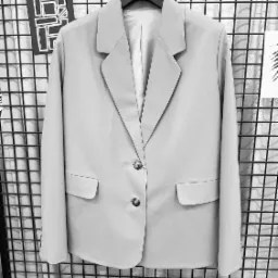 Jasnoszara marynarka damska, z dwoma guzikami, wisząca na wieszaku na tle metalowej siatki i abstrakcyjnych grafik.