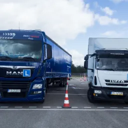 Dwa samochody ciężarowe z oznaczeniami kursu nauki jazdy, MAN z naczepą w kolorze niebieskim oraz IVECO w kolorze białym, stojące na placu manewrowym z pachołkami.