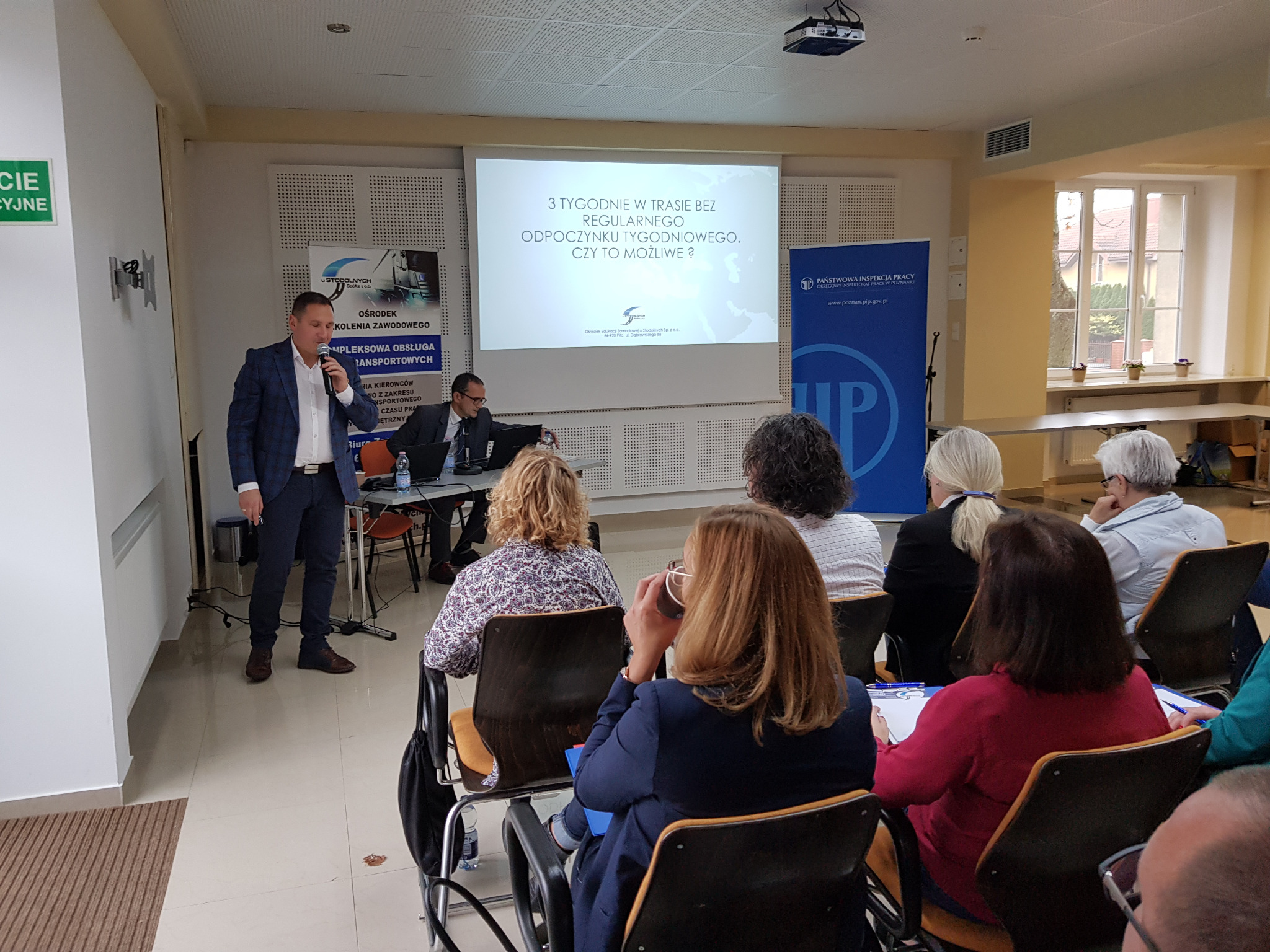 Prelegent w garniturze przemawia do grupy osób siedzących na krzesłach podczas prezentacji na temat czasu pracy kierowców, na ekranie wyświetlony tytuł '3 tygodnie w trasie bez regularnego...
