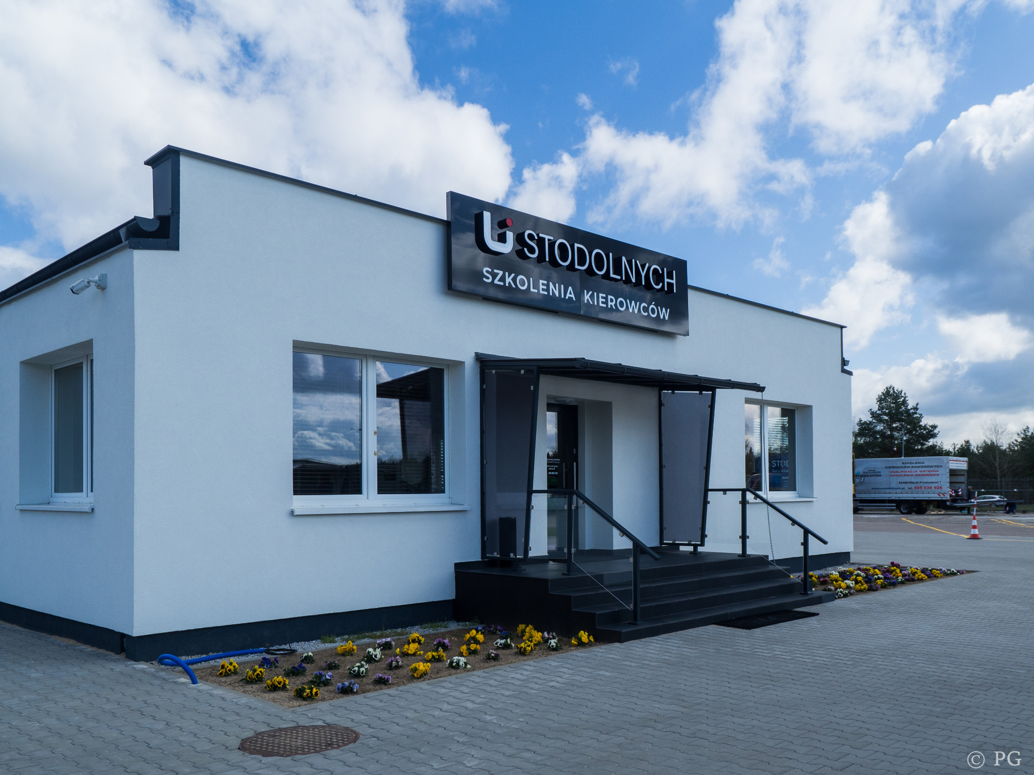 Biały budynek z szyldem 'U STODOLNYCH SZKOLENIA KIEROWCÓW', widoczne kwiaty przed wejściem i ciężarówka na placu manewrowym w tle.