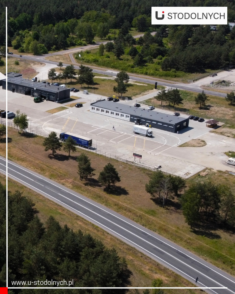 Widok z lotu ptaka na budynki z dużym parkingiem, na którym stoi kilka samochodów i ciężarówek, otoczone lasem i drogą.