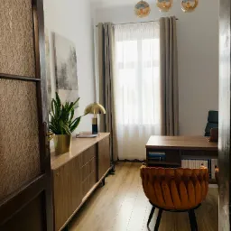 Wnętrze gabinetu z biurkiem, krzesłem w stylu retro, komodą z rośliną doniczkową i trzema wiszącymi lampami o kulistych kloszach.