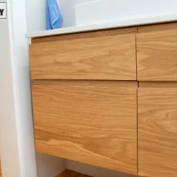 Drewniana szafka łazienkowa z czterema szufladami pod umywalką, widoczny fragment białego blatu i niebieski ręcznik, logo firmy w lewym górnym rogu.