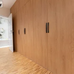 Długa, zabudowana szafa wnękowa z drewnopodobnymi frontami i czarnymi uchwytami na tle podłogi z desek oraz otwartych drzwi do pomieszczenia z tapetą w kwiaty.