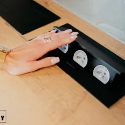 Blat drewniany z wbudowanym, czarnym panelem z trzema gniazdami elektrycznymi; dłoń kobiety z pierścionkami i złotym zegarkiem dotyka panelu.