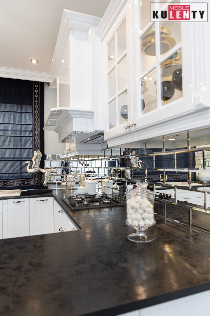 Elegancka kuchnia z białymi szafkami, lustrzanym backsplashiem i czarnym blatem, ozdobiona figurką żurawia i szklanym pojemnikiem z białymi kulkami.