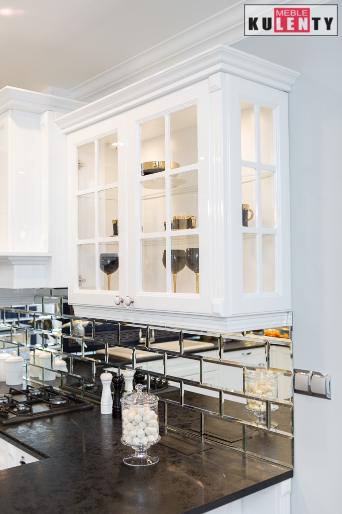 Biała, przeszklona szafka kuchenna z dekoracyjnymi elementami, zawieszona nad blatem z czarnego kamienia i lustrzanymi płytkami. Na blacie pojemnik ze słodyczami i przyprawy. W tle widoczne logo...
