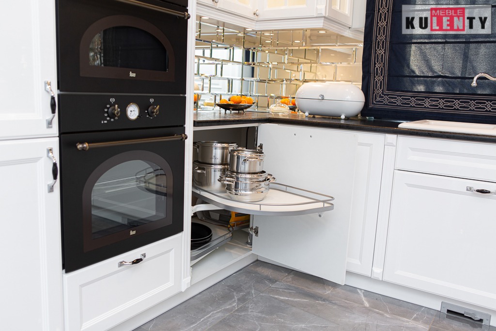 Biała kuchnia z zabudową, piekarnik w stylu retro, narożna szafka z wysuwanym systemem przechowywania garnków, szare płytki podłogowe.