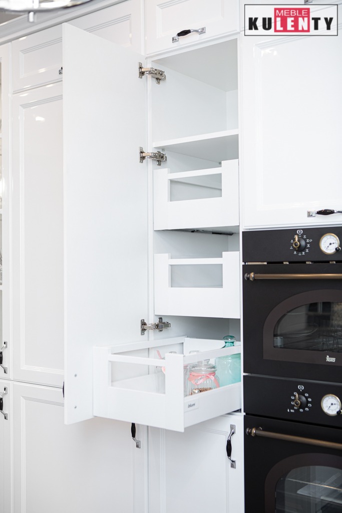 Biała szafka kuchenna z otwartymi drzwiami i wysuwanymi koszami; na dolnej półce szklane pojemniki z zawartością; obok czarny piekarnik z pokrętłami; widoczne zawiasy i uchwyty.