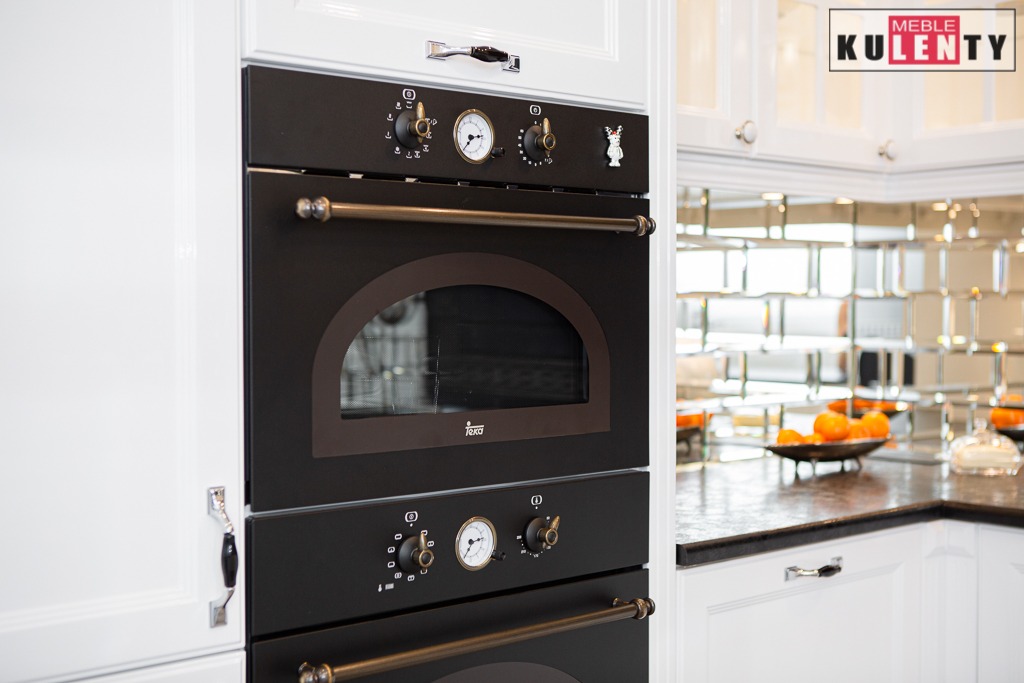 Zabudowa kuchenna z dwoma piekarnikami w stylu retro, czarne fronty, mosiężne uchwyty, białe szafki, mozaika lustrzana na ścianie, dekoracyjny magnes na piekarniku.